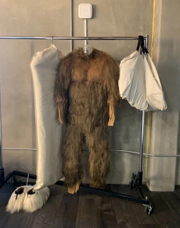 Furry brown costume hangs on a metal rack between two garment bags in front of a gray wall. The costume has fur-like material.